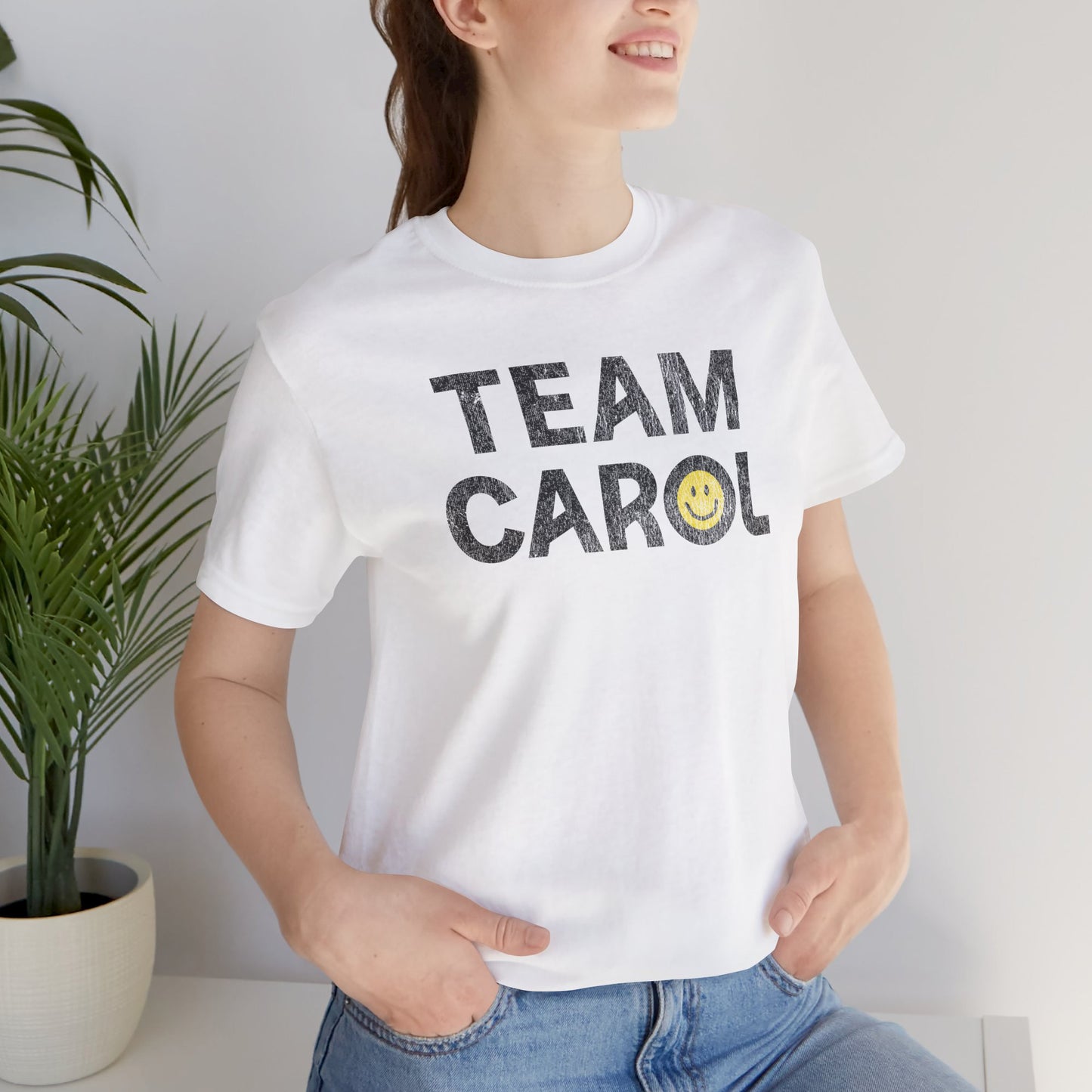 Person wearing white TEAM CAROL t-shirt inspired by Pluribus with vintage distressed smiley face graphic, seated near a plant