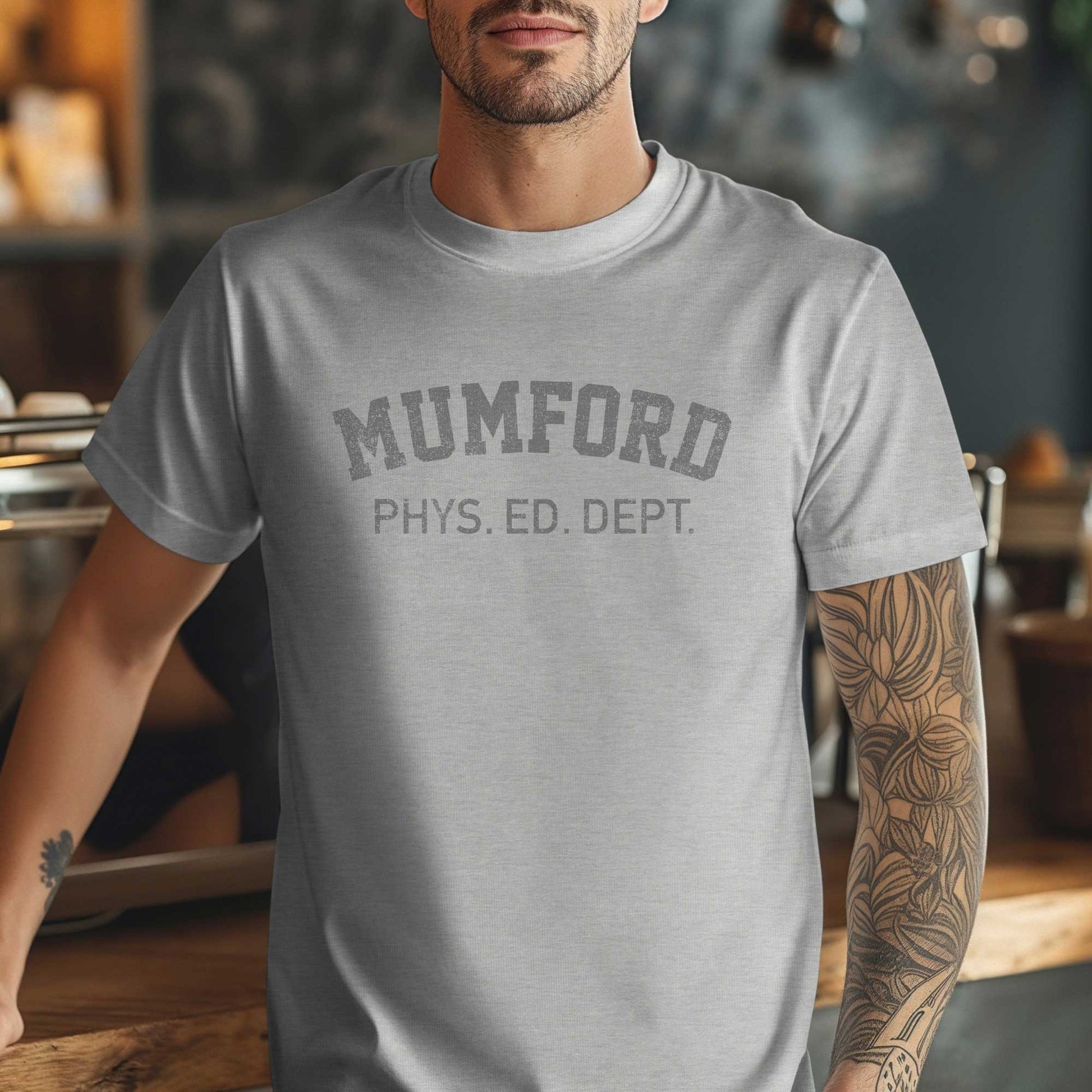 Male model wearing an Athletic Heather Grey Soft Bella+Canvas 3001 short-sleeve t-shirt gym tee featuring the Mumford Phys Ed logo graphic that Eddie Murphy wore as Axel Foley in the movie Beverly Hills Cop in a vintage distressed style, sold by ScriptedBrand.